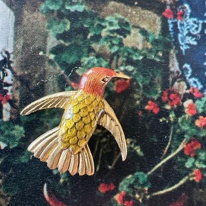 May include: A gold-colored hummingbird brooch with red and yellow enamel. The hummingbird is in flight with its wings spread out.