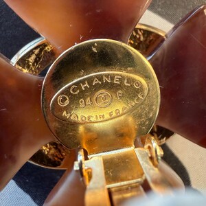 Puede incluir: Primer plano de un pendiente de clip dorado con dise&ntilde;o floral. El pendiente presenta un disco central dorado con el texto "CHANEL 94 P MADE IN FRANCE". Los p&eacute;talos de la flor son de color marr&oacute;n transl&uacute;cido.