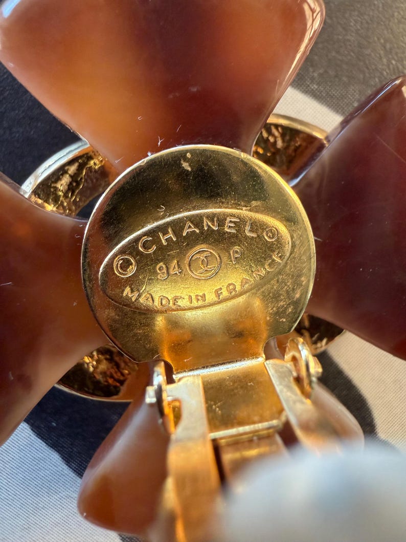 Puede incluir: Primer plano de un pendiente de clip dorado con dise&ntilde;o floral. El pendiente presenta un disco central dorado con el texto "CHANEL 94 P MADE IN FRANCE". Los p&eacute;talos de la flor son de color marr&oacute;n transl&uacute;cido.