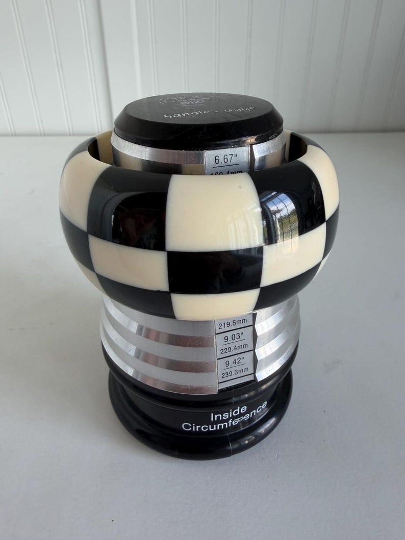 May include: A black and white checkered bangle bracelet displayed on a tiered sizing mandrel. The mandrel features printed measurements including 6.67 inches, 9.03 inches, and 9.42 inches, with the text Inside Circumference printed at the base.