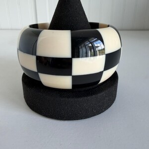 May include: A wide bangle bracelet featuring a high-gloss finish with a repeating black and white checkerboard pattern. The bracelet has a smooth, rounded exterior surface and a thick, solid construction that maintains a consistent circular shape.