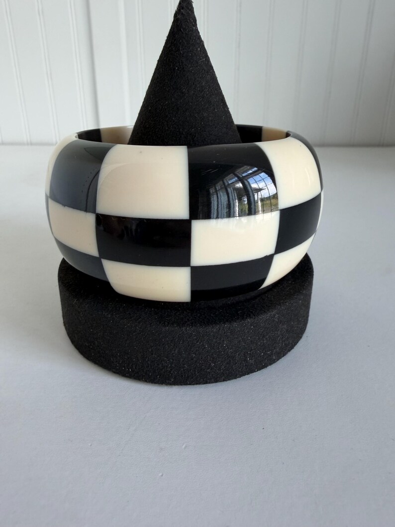 May include: A wide bangle bracelet featuring a high-gloss finish with a repeating black and white checkerboard pattern. The bracelet has a smooth, rounded exterior surface and a thick, solid construction that maintains a consistent circular shape.