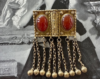 May include: A vintage gold-toned brooch with two oval, red-colored stones. The brooch has a rectangular frame with intricate detailing and dangling chains ending in small, round beads. The background features a black and white image of a woman.