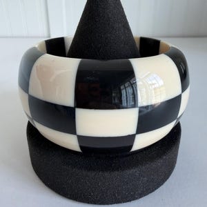 May include: A wide bangle bracelet featuring a high-gloss finish with a repeating black and off-white checkerboard pattern. The bracelet has a smooth, rounded exterior surface and a consistent width throughout its circular design.