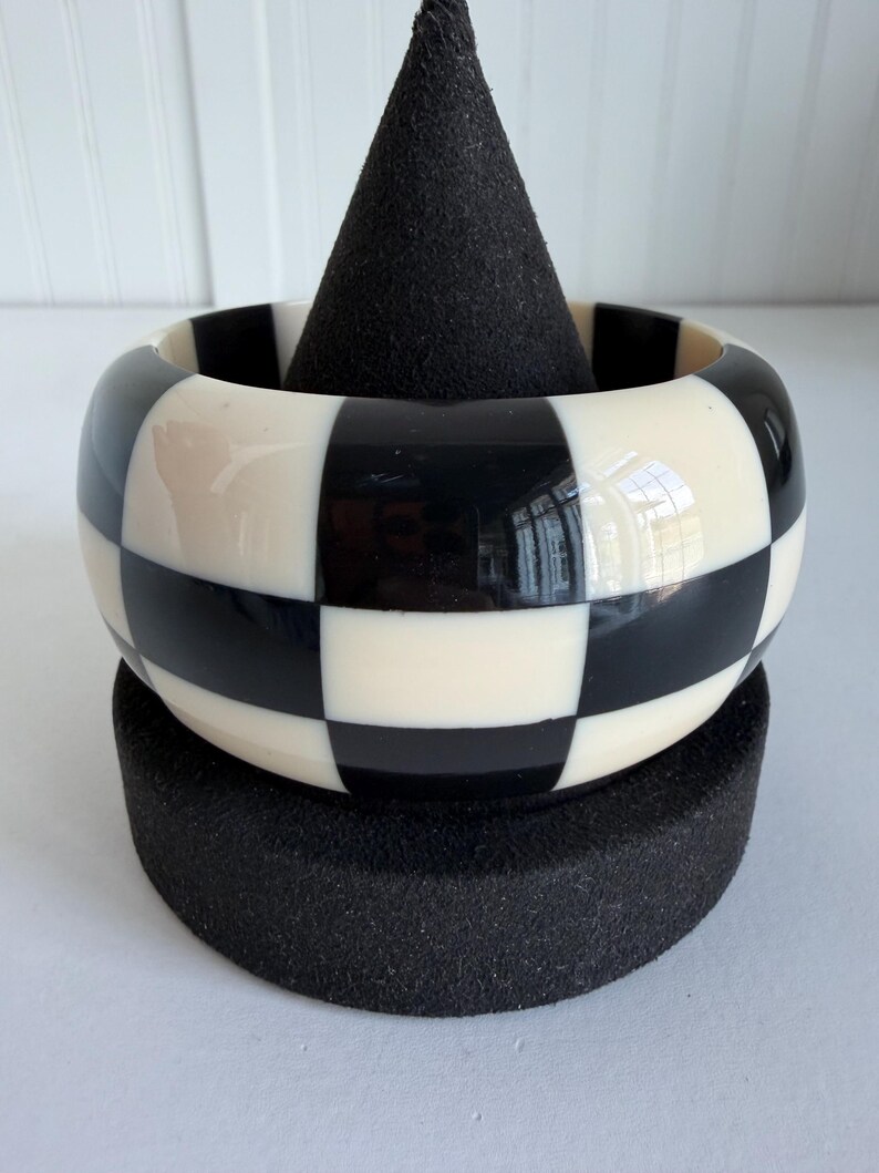 May include: A wide bangle bracelet featuring a high-gloss finish with a repeating black and off-white checkerboard pattern. The bracelet has a smooth, rounded exterior surface and a consistent width throughout its circular design.