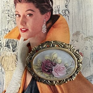 May include: A gold-toned oval brooch with a cameo-like design featuring two roses, one pink and one burgundy, with green leaves. The brooch has a detailed ornate frame.