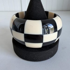 May include: A wide bangle bracelet featuring a high-contrast, repeating checkerboard pattern of alternating black and off-white square tiles. The surface of the bracelet is smooth and glossy, reflecting light across its rounded, circular form.