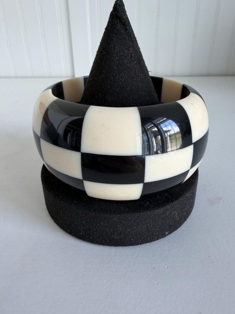 May include: A wide bangle bracelet featuring a high-contrast, repeating checkerboard pattern of alternating black and off-white square tiles. The surface of the bracelet is smooth and glossy, reflecting light across its rounded, circular form.