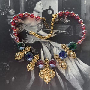 May include: A vintage necklace featuring a gold chain and ornate gold pendants. The necklace has red and clear glass beads, and the pendants hold blue, green, and red glass stones. The necklace is displayed against a black and white background.