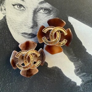 Puede incluir: Un par de pendientes marrones y dorados con forma de flor. Cada pendiente presenta un logotipo doble C dorado en el centro. Los pendientes tienen un patr&oacute;n de carey y est&aacute;n colocados sobre un fondo fotogr&aacute;fico vintage en blanco y negro.
