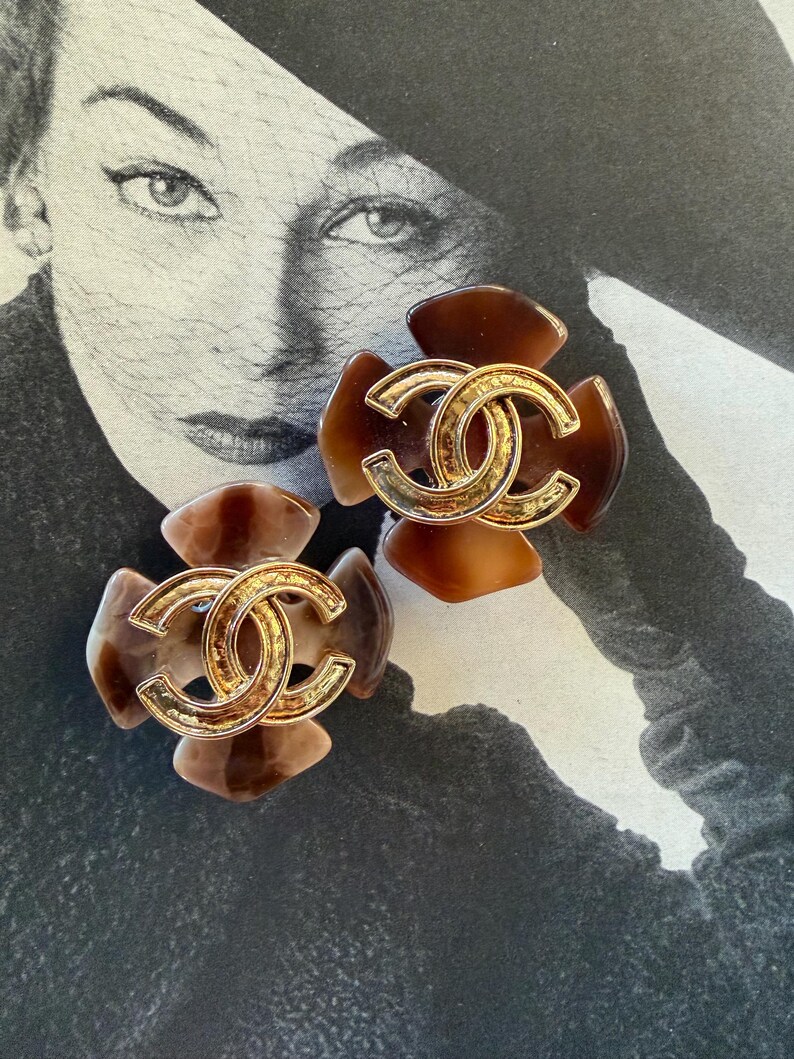 Puede incluir: Un par de pendientes marrones y dorados con forma de flor. Cada pendiente presenta un logotipo doble C dorado en el centro. Los pendientes tienen un patr&oacute;n de carey y est&aacute;n colocados sobre un fondo fotogr&aacute;fico vintage en blanco y negro.