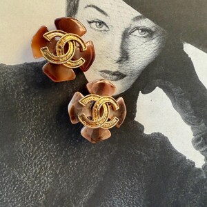 Puede incluir: Dos pendientes con forma de flor con el logotipo CC entrelazado en tono dorado en el centro. Los p&eacute;talos presentan tonos marrones y beige. Los pendientes est&aacute;n colocados sobre una fotograf&iacute;a vintage en blanco y negro de una mujer.