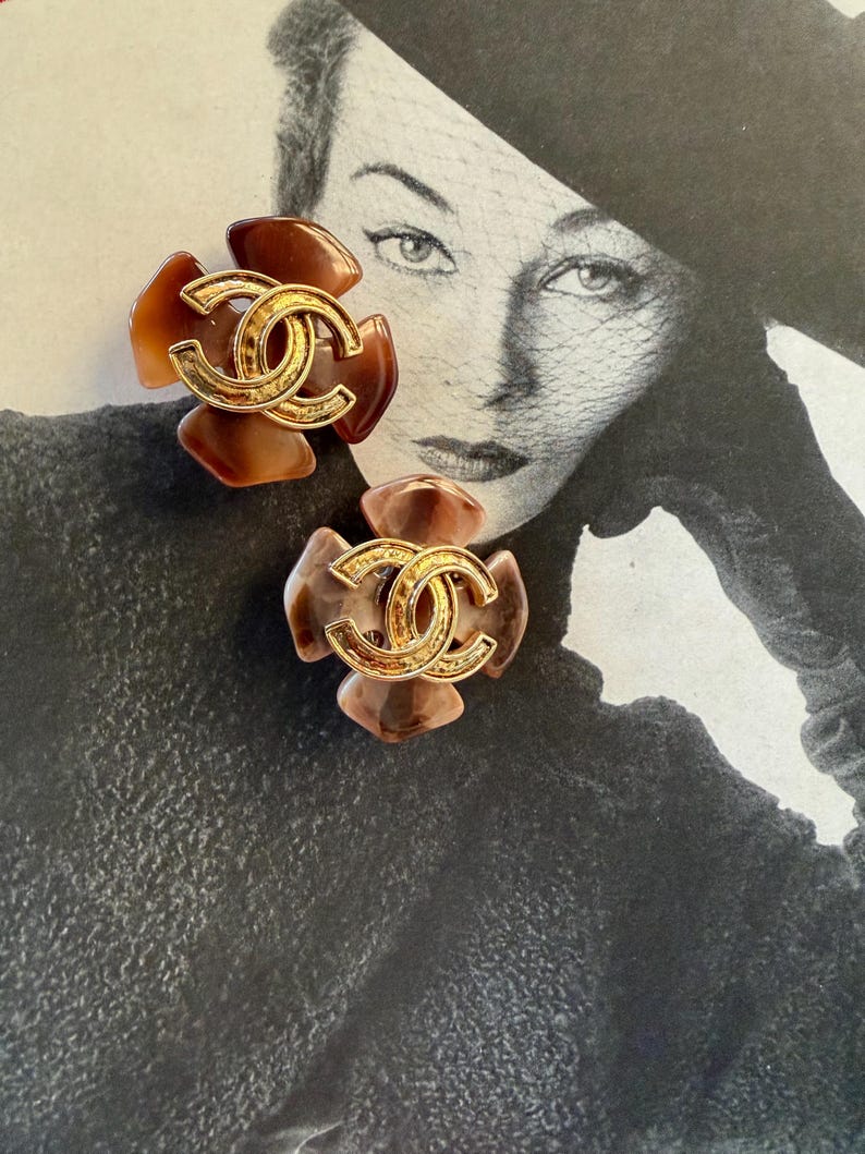 Puede incluir: Dos pendientes con forma de flor con el logotipo CC entrelazado en tono dorado en el centro. Los p&eacute;talos presentan tonos marrones y beige. Los pendientes est&aacute;n colocados sobre una fotograf&iacute;a vintage en blanco y negro de una mujer.