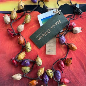 May include: A long necklace featuring a dark brown crocheted cord adorned with numerous small, multi-colored seed pods in shades of red, yellow, orange, purple, and beige. A large, rectangular green tag with the text Maria Oiticica is attached.