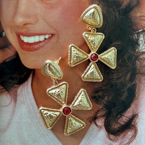 May include: A pair of gold-toned dangle earrings featuring a cross-shaped design composed of four hammered triangular metal pieces. Each earring is centered with a small, round, deep red stone and includes a triangular stud top piece.