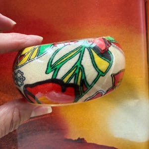 May include: A wide bangle bracelet featuring a cream-colored fabric base with a vibrant floral pattern. The design includes bold red, yellow, and green stylized flowers and leaves with black outlines, all encased in a smooth, glossy, clear finish.