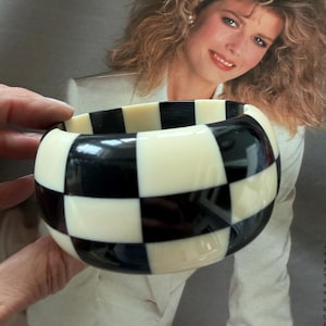 May include: A wide bangle bracelet featuring a high-contrast checkerboard pattern of alternating glossy black and off-white square tiles. The circular accessory has a smooth, polished finish and a thick, rounded profile that creates a bold geometric look.