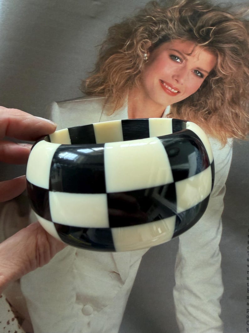 May include: A wide bangle bracelet featuring a high-contrast checkerboard pattern of alternating glossy black and off-white square tiles. The circular accessory has a smooth, polished finish and a thick, rounded profile that creates a bold geometric look.