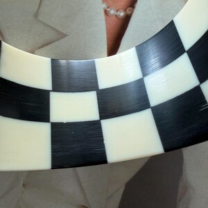 May include: A wide, curved bangle bracelet featuring a high-contrast checkerboard pattern of alternating black and off-white squares. The surface of the bracelet has a smooth, polished finish with visible fine linear textures throughout the material.