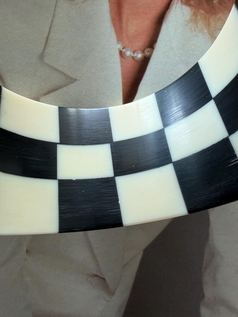 May include: A wide, curved bangle bracelet featuring a high-contrast checkerboard pattern of alternating black and off-white squares. The surface of the bracelet has a smooth, polished finish with visible fine linear textures throughout the material.