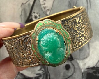 May include: Vintage gold-tone hinged bracelet with an ornate floral design. The bracelet features a central green cameo set in a green and gold frame. The bracelet is approximately 1 inch wide.