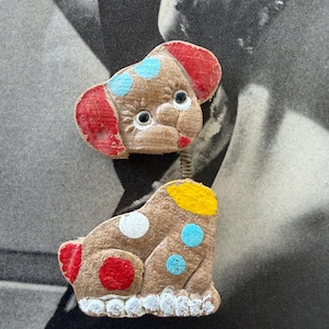 May include: Two vintage dog-shaped brooches. The top brooch is a dog head with red ears and blue spots. The bottom brooch is a dog body with red, white, and blue spots. Both are brown with white trim.