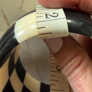 May include: A black and white checkered bangle bracelet featuring a smooth, glossy finish. A white measuring tape is wrapped around the width of the bangle, clearly indicating a measurement of approximately 2 inches across the band's surface.