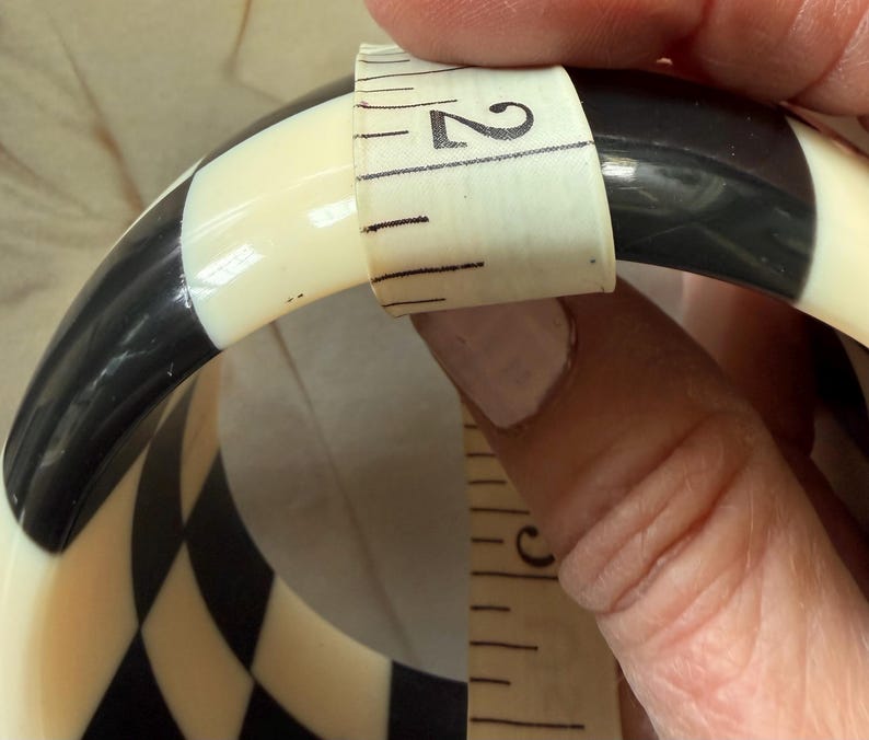 May include: A black and white checkered bangle bracelet featuring a smooth, glossy finish. A white measuring tape is wrapped around the width of the bangle, clearly indicating a measurement of approximately 2 inches across the band's surface.