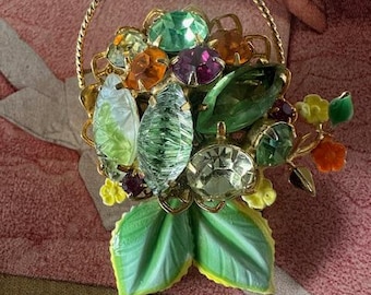 May include: A gold tone brooch with a basket filled with green, orange, and purple rhinestones. The basket is surrounded by green leaves.