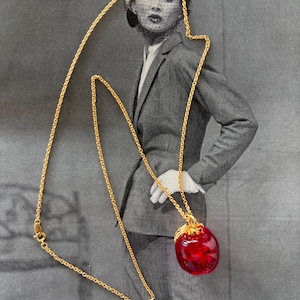 May include: A gold-toned necklace with a large, red, apple-shaped pendant. The pendant has a textured surface and is suspended from a delicate chain. The background is a vintage black and white photograph of a woman in a suit.