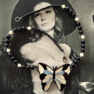 May include: A black beaded necklace with a butterfly pendant. The pendant features a black, white, and light blue butterfly design. The necklace has a silver clasp and is set against a black and white background.