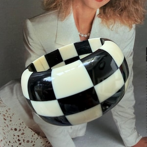 May include: A wide, chunky bangle bracelet featuring a high-contrast, repeating checkerboard pattern in solid black and off-white squares. The bracelet has a smooth, rounded surface and a thick, substantial profile that encircles the wrist.