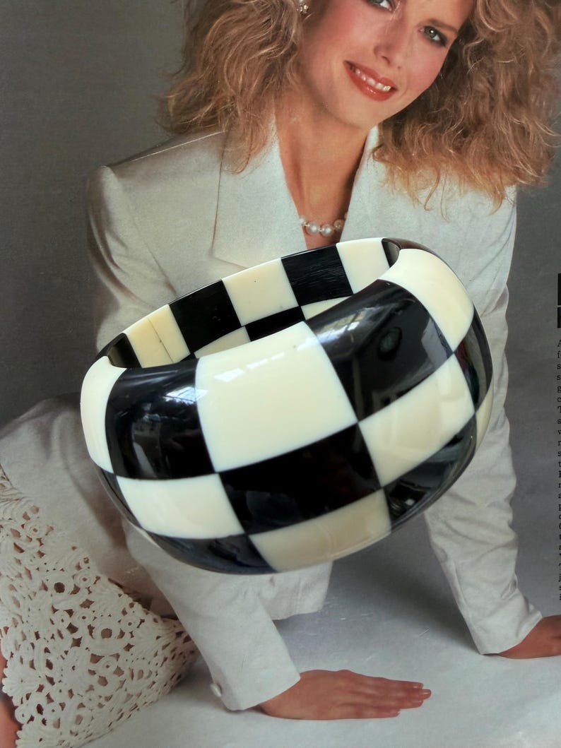May include: A wide, chunky bangle bracelet featuring a high-contrast, repeating checkerboard pattern in solid black and off-white squares. The bracelet has a smooth, rounded surface and a thick, substantial profile that encircles the wrist.