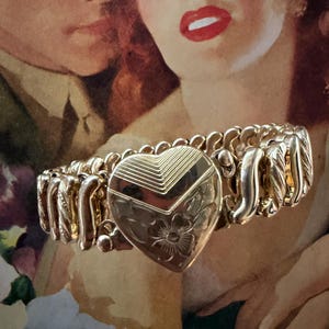 May include: A gold-toned bracelet with a heart-shaped centerpiece. The heart features engraved floral and geometric designs. The bracelet has an expandable, stretchable design. The background features a vintage illustration of a couple.