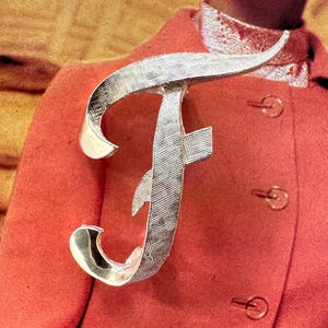 May include: A silver-toned letter "F" brooch with a textured surface. The brooch is pinned to a red coat. The brooch is a decorative accessory.