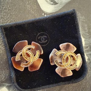 Puede incluir: Un par de pendientes vintage de clip con un dise&ntilde;o floral y un logotipo CC entrelazado en tono dorado. Los pendientes tienen un patr&oacute;n de carey y se muestran en una bolsa de terciopelo negro.