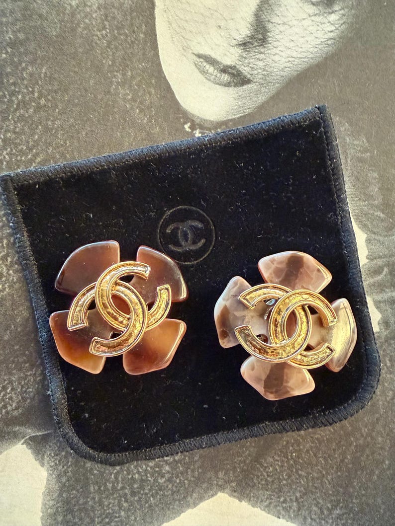 Puede incluir: Un par de pendientes vintage de clip con un dise&ntilde;o floral y un logotipo CC entrelazado en tono dorado. Los pendientes tienen un patr&oacute;n de carey y se muestran en una bolsa de terciopelo negro.