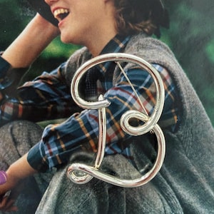 May include: A silver-toned metal brooch in the shape of the letter "B". The brooch has a cursive, stylized design with a pin on the back. The background features a person wearing a plaid shirt and a hat.