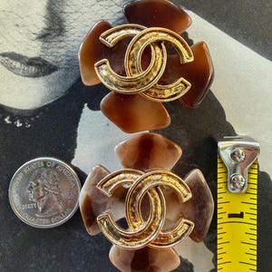 Puede incluir: Par de pendientes vintage marrones y dorados. Cada pendiente presenta un logotipo CC entrelazado dorado en el centro, rodeado de detalles marrones en forma de p&eacute;talo. Los pendientes tienen aproximadamente 3,8 cm de di&aacute;metro.