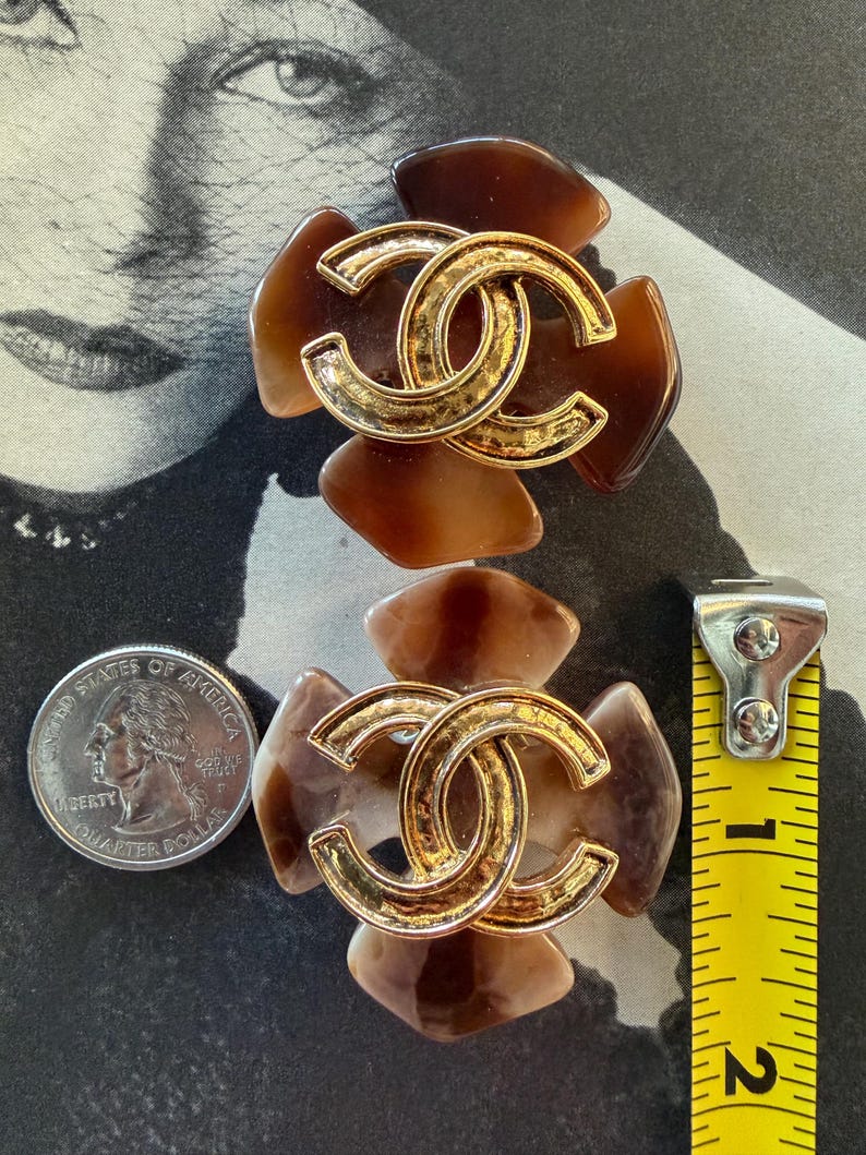 Puede incluir: Par de pendientes vintage marrones y dorados. Cada pendiente presenta un logotipo CC entrelazado dorado en el centro, rodeado de detalles marrones en forma de p&eacute;talo. Los pendientes tienen aproximadamente 3,8 cm de di&aacute;metro.
