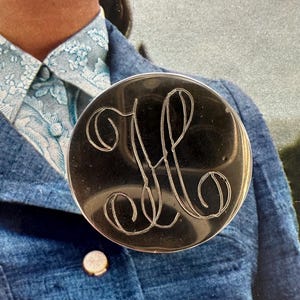 May include: A round, silver-toned brooch with an engraved monogram of the letters "M" and "P" in a cursive script. The brooch is set against a blue jacket with a gold button.