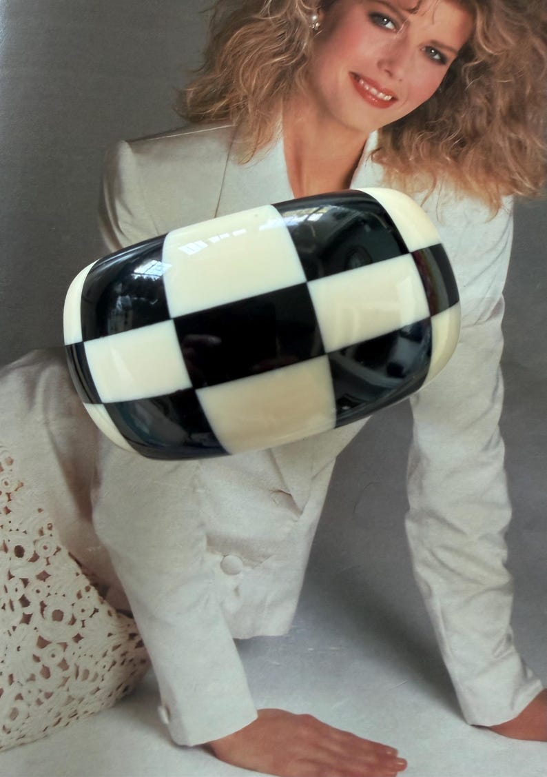 May include: A wide, chunky bangle bracelet featuring a high-gloss finish with a bold, repeating black and white checkerboard pattern. The bracelet has a rounded, thick profile and a smooth, polished surface that reflects light across its surface.