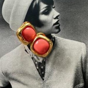 May include: Gold-tone clip-on earrings with a square, coral-colored center. The earrings have a thick, textured gold border. The earrings are shown against a black and white background of a woman wearing a hat and a collared shirt.