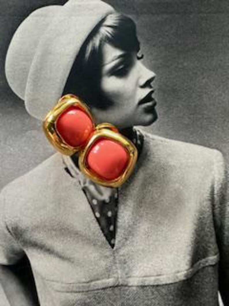 May include: Gold-tone clip-on earrings with a square, coral-colored center. The earrings have a thick, textured gold border. The earrings are shown against a black and white background of a woman wearing a hat and a collared shirt.