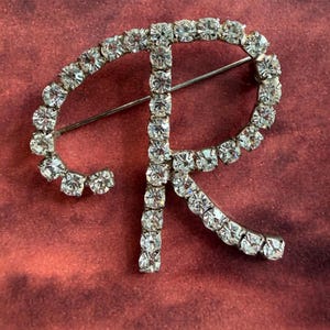 May include: A silver-toned metal brooch in the shape of the uppercase letter R, featuring a continuous row of clear, round-cut rhinestones set along the entire surface of the letter. A thin metal pin mechanism is visible on the back of the brooch.