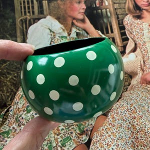 May include: A round, glossy green bowl featuring a pattern of white polka dots. The interior of the bowl is black, and the exterior surface is smooth with a rounded shape. The bowl is held in place, showcasing its vibrant green color and white spots.