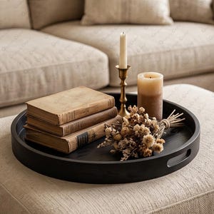 Peut inclure: Un plateau rond en bois noir avec des poignées intégrées contenant une pile de trois livres anciens à couverture rigide, une longue bougie fine blanche dans un bougeoir en laiton, une bougie pilier beige épaisse et un bouquet de fleurs séchées.