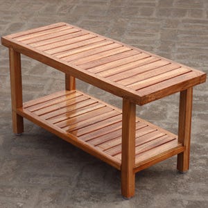 Puede incluir: Banco de madera con tapa de listones y estante inferior. El banco es de madera marrón claro y tiene forma rectangular. El banco mide aproximadamente 122 cm de largo, 41 cm de ancho y 46 cm de alto.