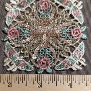 May include: A delicate lace applique with a floral design featuring pink and green embroidery. The applique is adorned with gold sequins and measures approximately 3 inches in diameter.