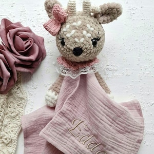 Könnte beinhalten: Ein gehäkeltes Stofftier in Form eines Rehs mit einer rosa Schleife und einem rosa-weißen Spitzenkragen. Das Reh hält eine rosa Musselin-Babydecke mit dem Namen "Eddle" bestickt.
